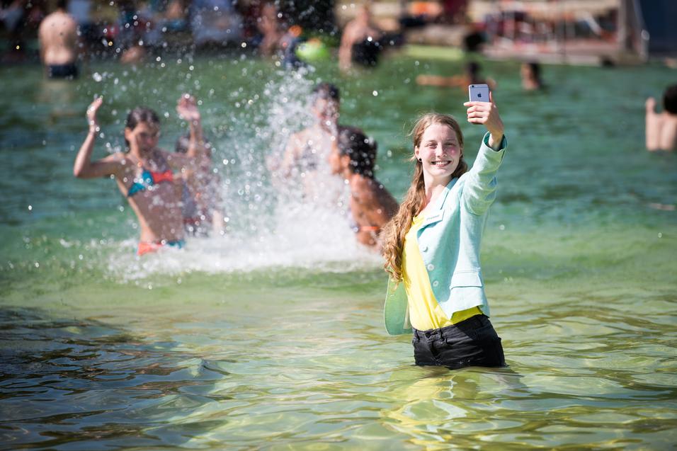 Lächelnde Frau im Wasser, die ein Selfie macht, während Kinder im Hintergrund spielen und spritzen.