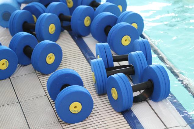 Blaue Hanteln aus Schaumstoff liegen auf einem Tisch an einem Schwimmbeckenrand.