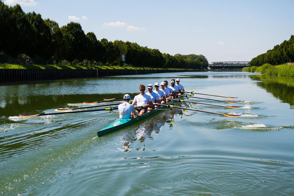 Acht Ruderer in weißen Shirts rudern synchron auf einem ruhigen Gewässer, umgeben von Bäumen und einem blauen Himmel.