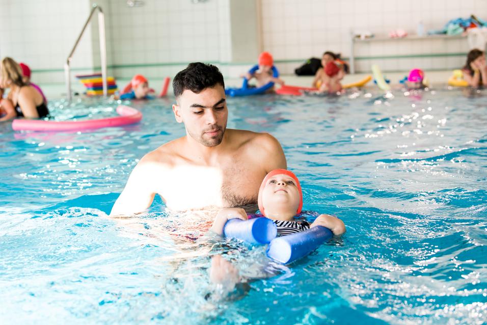Mann unterstützt ein Kind mit Schwimmhilfen im Wasser eines Schwimmbads, während andere Schwimmer im Hintergrund sind.