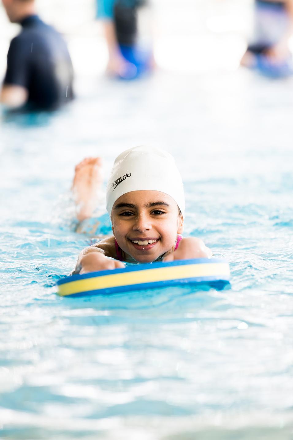 Lächelndes Mädchen schwimmt im Wasser auf einer Schwimmbrett, umgeben von anderen Schwimmern.